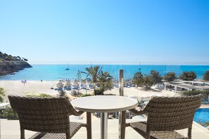 A balcony with wicker chairs and a table overlooking a beach with umbrellas and a pool. - Universal Hotel Castell Royal (Capdepera)
