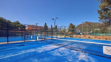 Una cancha de tenis al aire libre con una red, rodeada por una cerca, y un cielo azul despejado arriba.