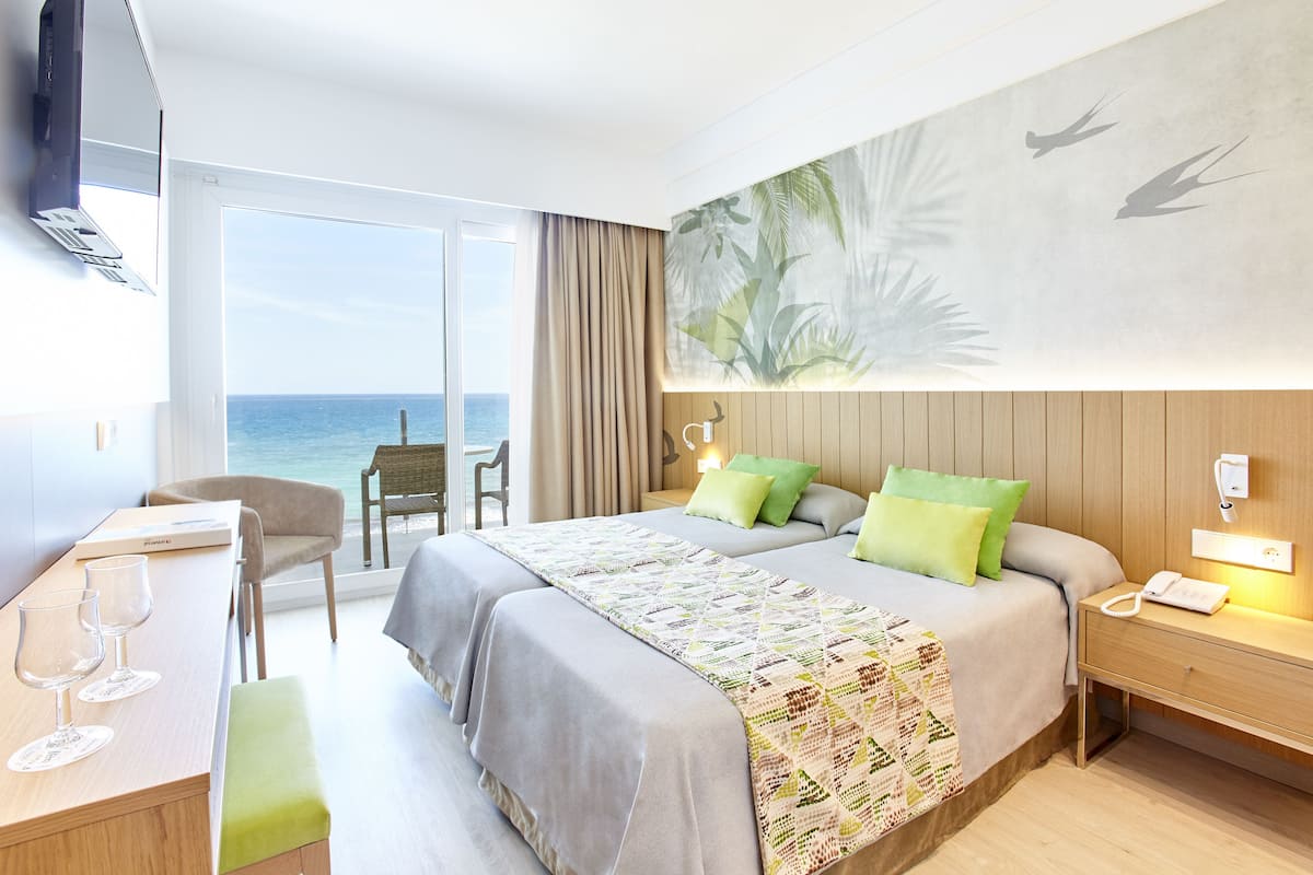 A hotel room with a bed, a desk with glasses, a chair, a nightstand, and a view of the ocean.
