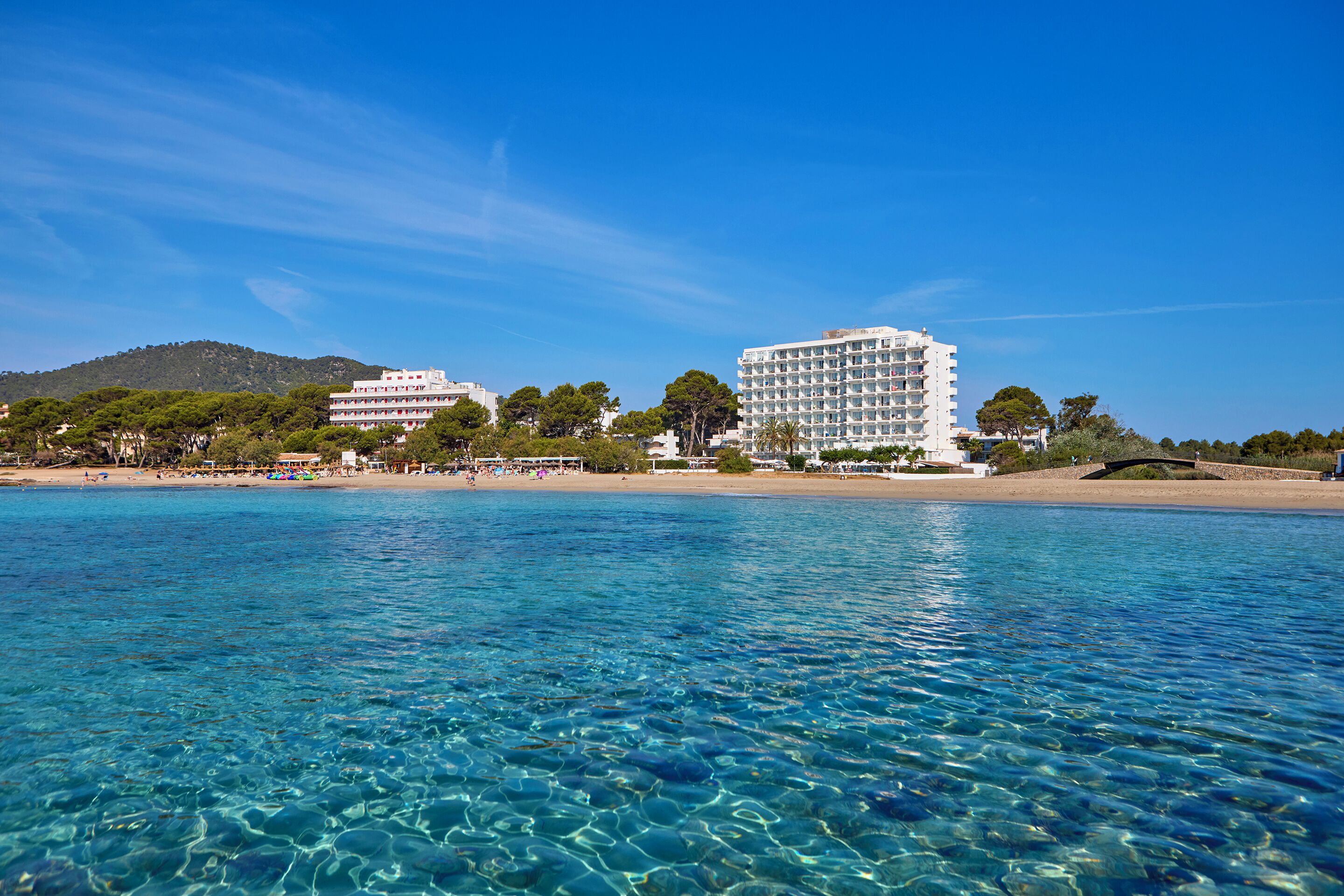 A coastal hotel with clear blue waters, a beach, and a mountain in the background.