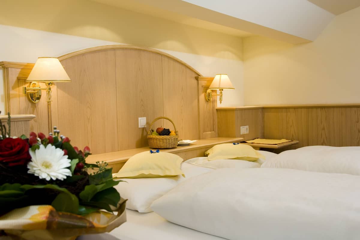 A hotel room with two beds, a wooden headboard, two wall-mounted lamps, a bedside table with a basket of fruit, and a floral arrangement.
