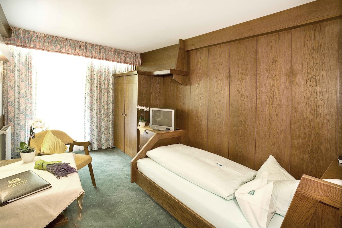 A compact bedroom with a wooden bed, a desk with a laptop, a chair, and a television on a wooden stand.