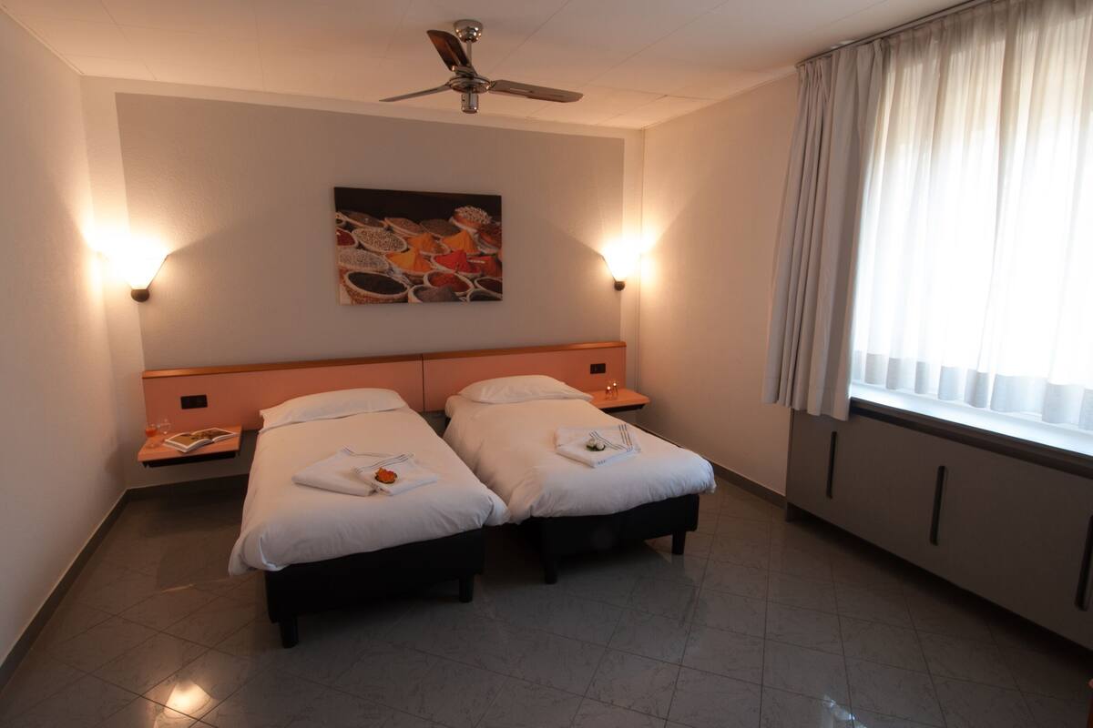 A hotel room with two single beds, a ceiling fan, a painting on the wall, and a window with curtains.