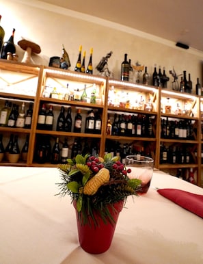 A table with a red vase containing a decorative arrangement of greenery, berries, and a corn cob. In the background, there are shelves with various bottles of wine. - Bahnhof Haus (Chiasso)