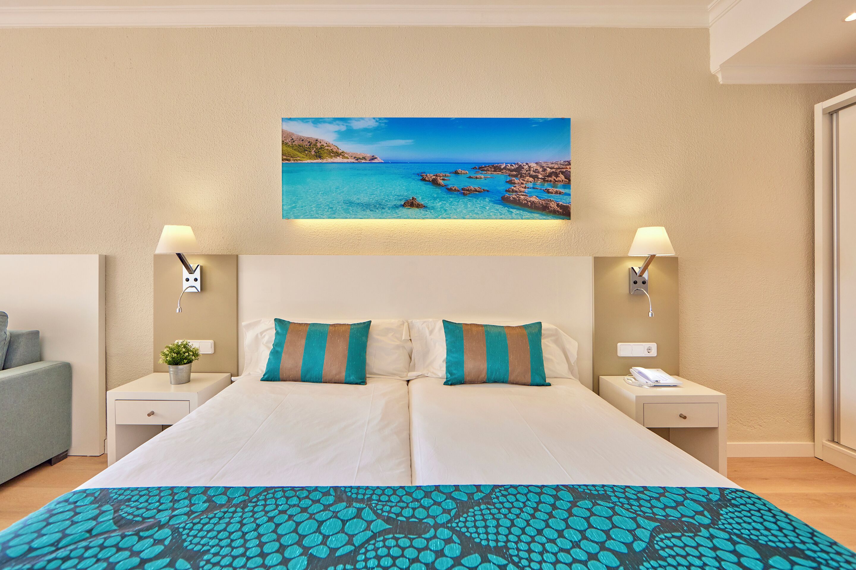 Habitación de hotel con una cama, dos mesitas de noche, una lámpara y un cuadro de una escena costera.