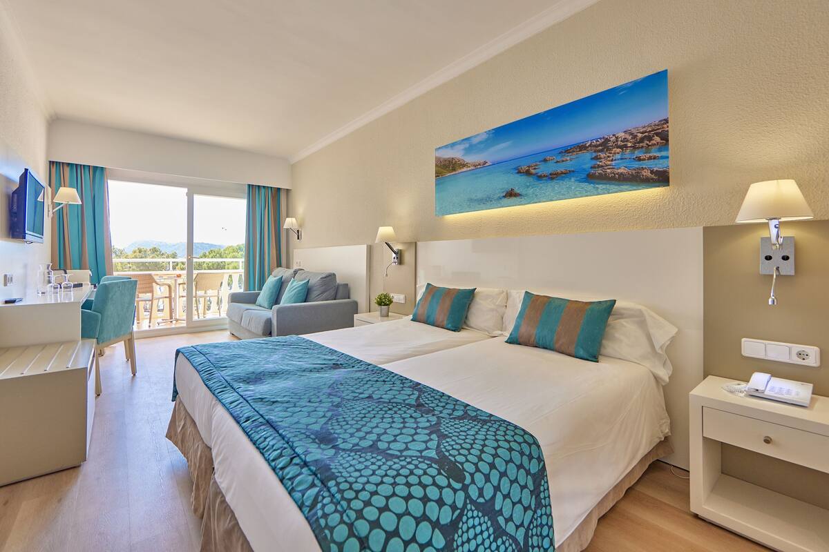 A hotel room with a large bed, a desk, a chair, and a balcony with a view of the sea.
