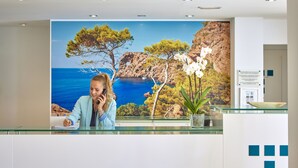 A reception desk with a scenic coastal mural, a potted orchid, and a glass-top counter. - Universal Hotel Lido Park & Spa (Calvia)