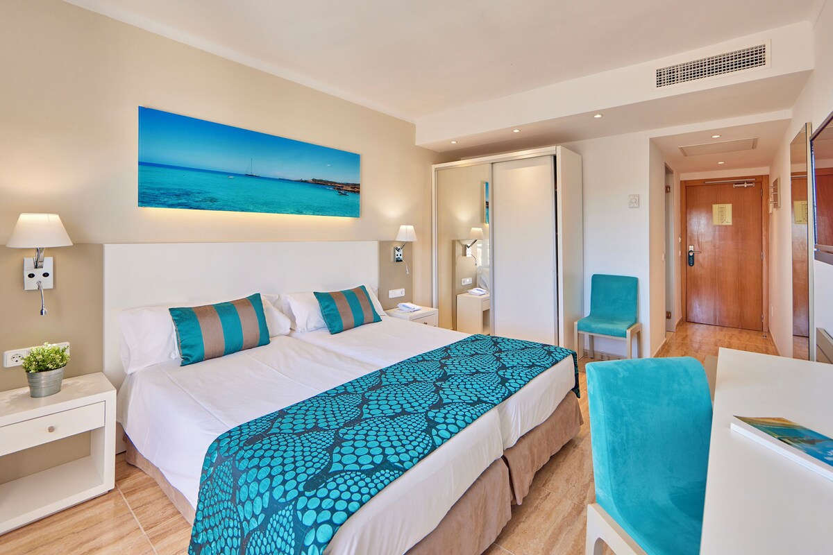 A hotel room with a bed, bedside lamps, a nightstand, a chair, a desk, and a large window.