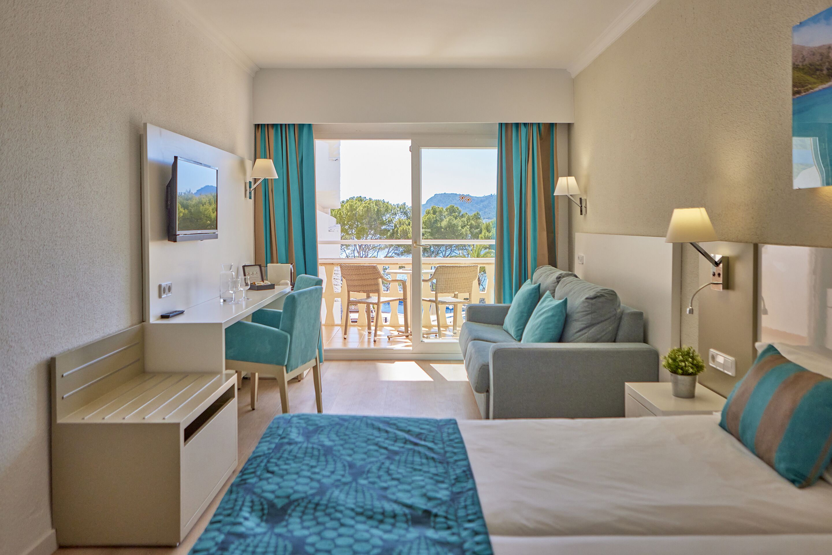 A hotel room with a bed, a desk, a chair, a sofa, and a balcony with a view of the mountains and trees.