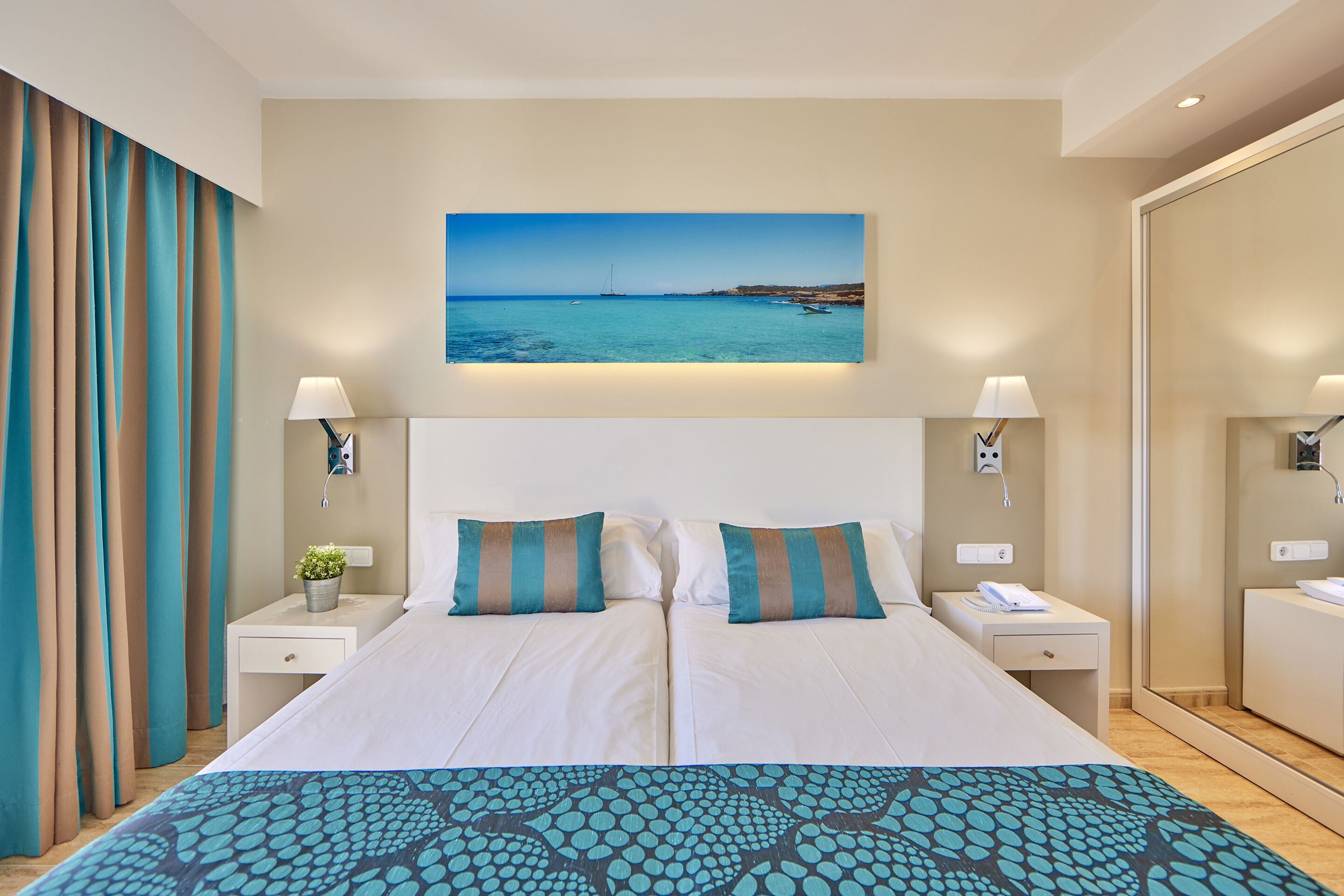 A double bed with a patterned bedspread, two pillows, a nightstand with a lamp, a small plant, and a painting of a coastal scene above the bed.