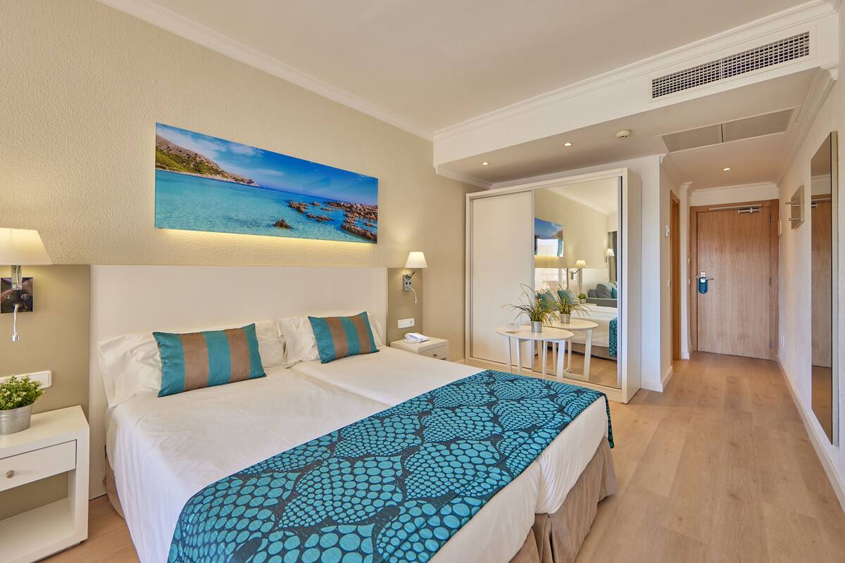 A hotel room with a bed, a nightstand, a lamp, a painting of a beach, and a hallway leading to a bathroom.