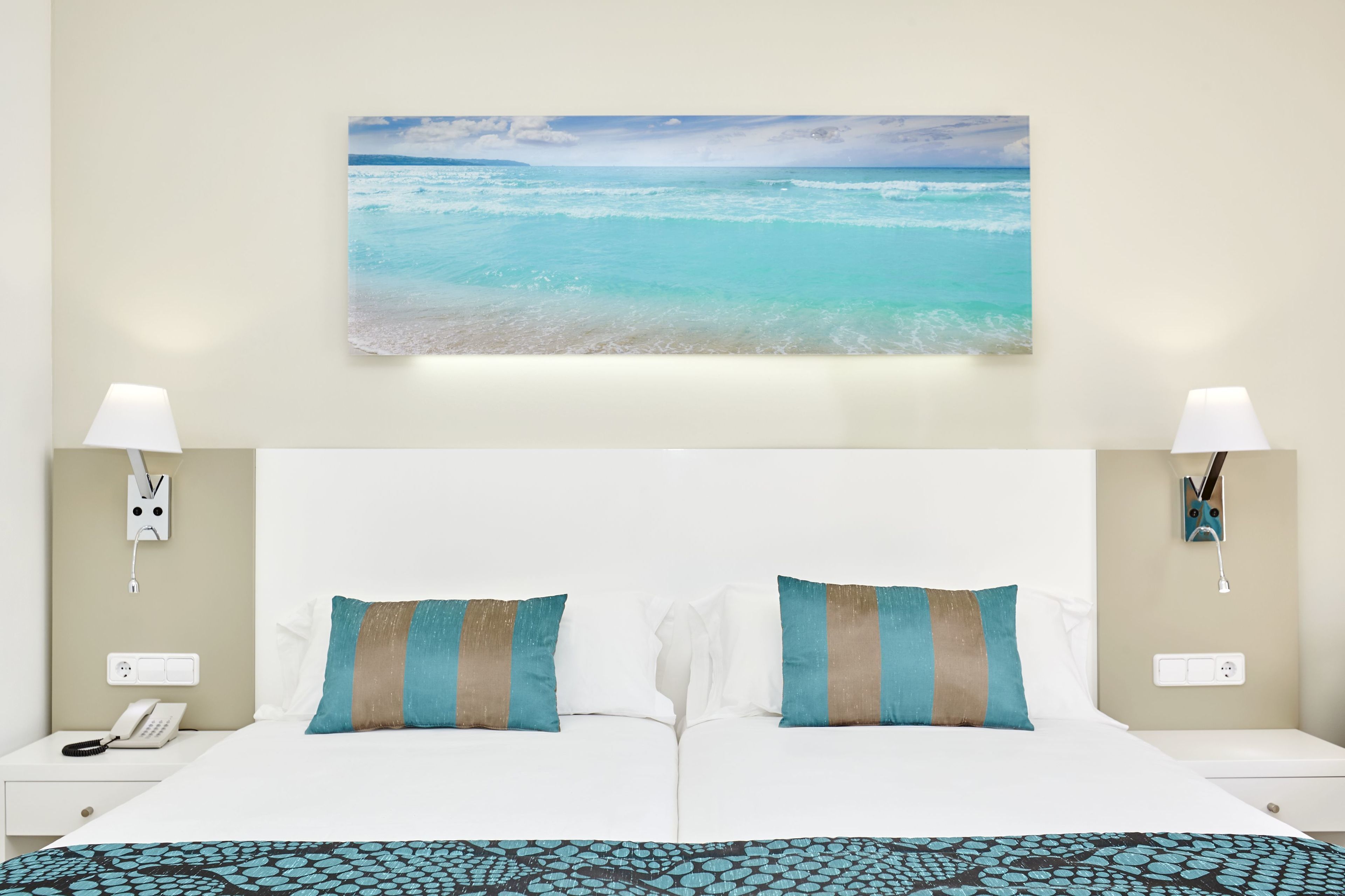 Habitación de hotel con una cama grande, dos lámparas de noche, un teléfono en la mesita y un cuadro de una playa en la pared.