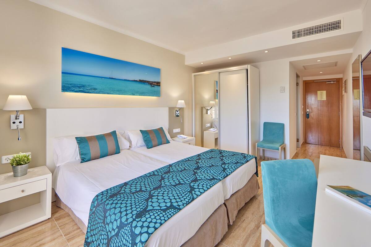 A hotel room with a bed, bedside lamps, a nightstand, a chair, a desk, and a large window.