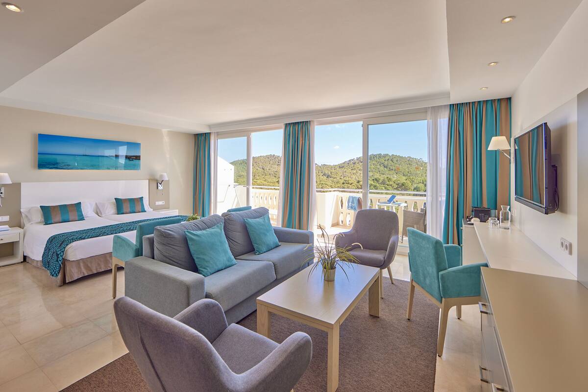A modern hotel room with a bed, sofa, chairs, a coffee table, and a TV.