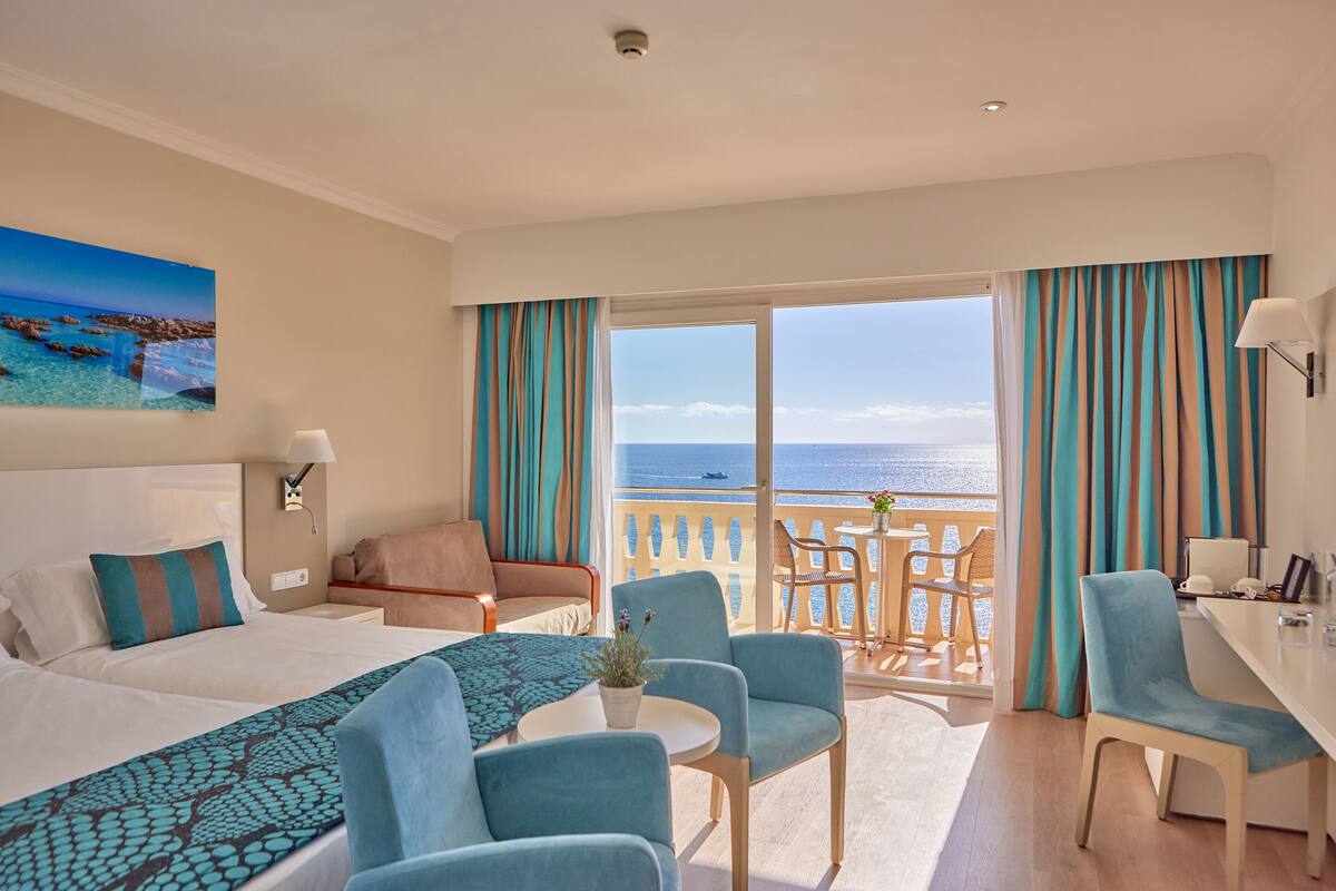 A hotel room with a bed, two chairs, a desk, and a balcony with a view of the sea.