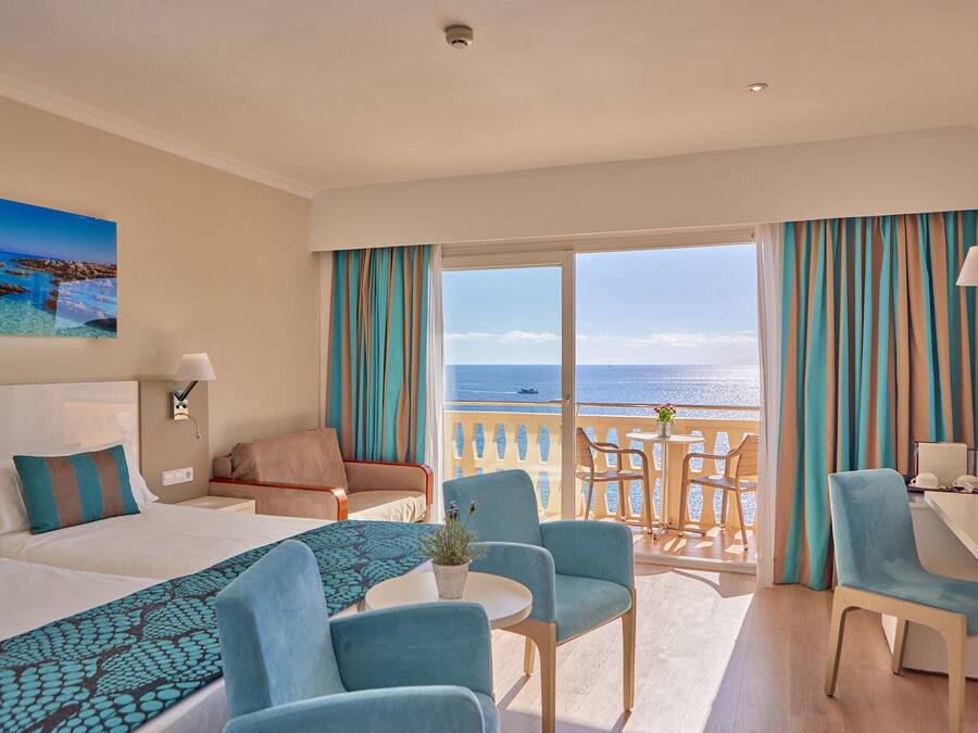 Ein Hotelzimmer mit Bett, zwei Stühlen, einem Schreibtisch und einem Balkon mit Meerblick.