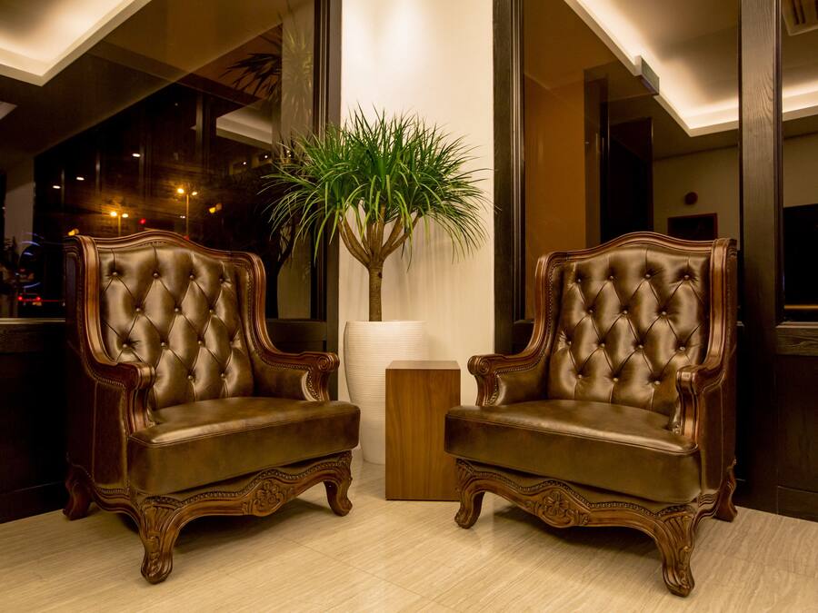 Deux fauteuils en cuir à accoudoirs, dotés d’un capitonnage et de pieds en bois sculptés, sont placés devant un grand vase blanc dans lequel se trouve une plante verte.