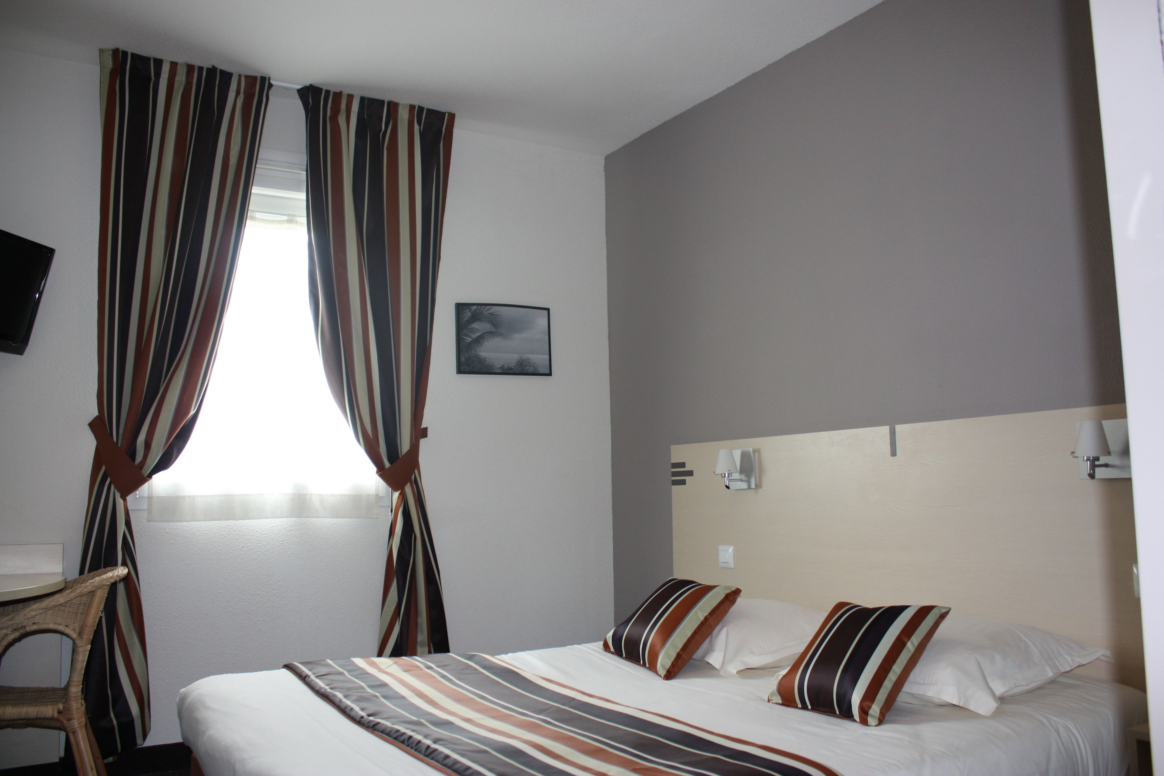 Una habitación de hotel con cama, cortinas, una silla, un televisor de pantalla plana y una lámpara de pared.