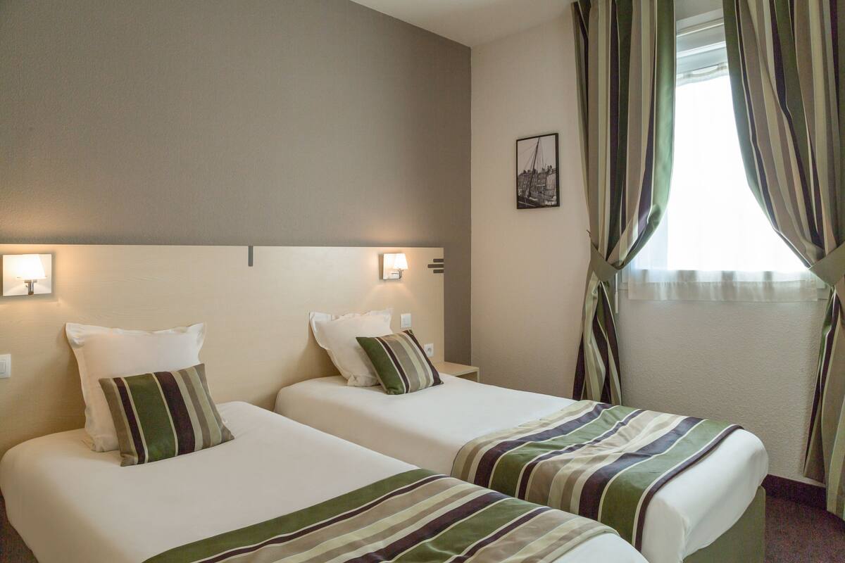 Une chambre d’hôtel avec deux lits simples, chacun équipé d’un oreiller et d’une couverture à rayures. Il y a une table de chevet avec une lampe entre les lits, ainsi qu’une fenêtre avec des rideaux.