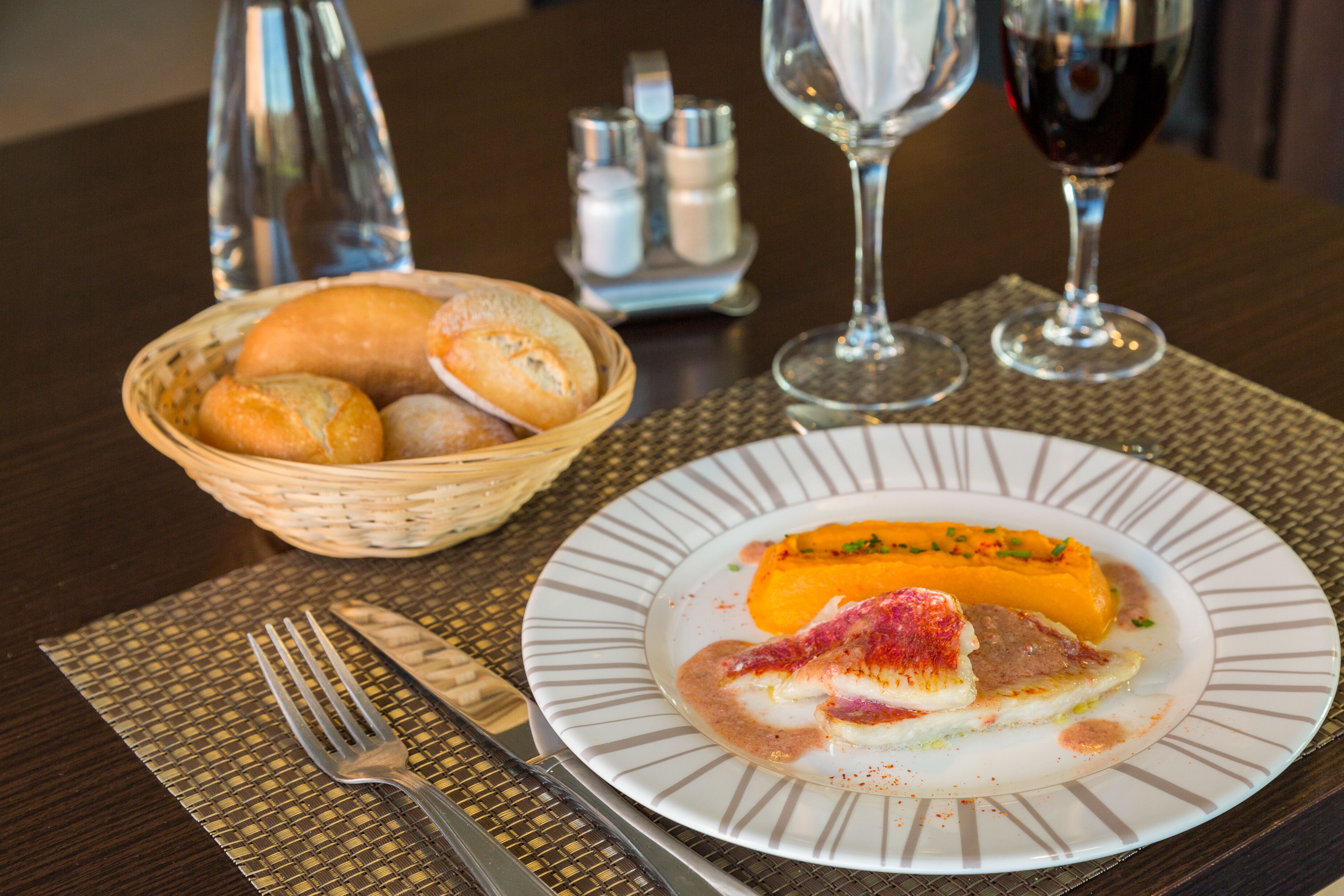 A dining table with a plate of food, a basket of bread, a wine glass, and a water bottle.