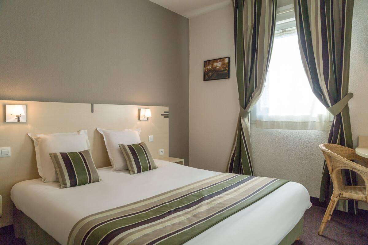 Une chambre d’hôtel avec un lit, deux lampes de chevet, une chaise et une fenêtre avec des rideaux.