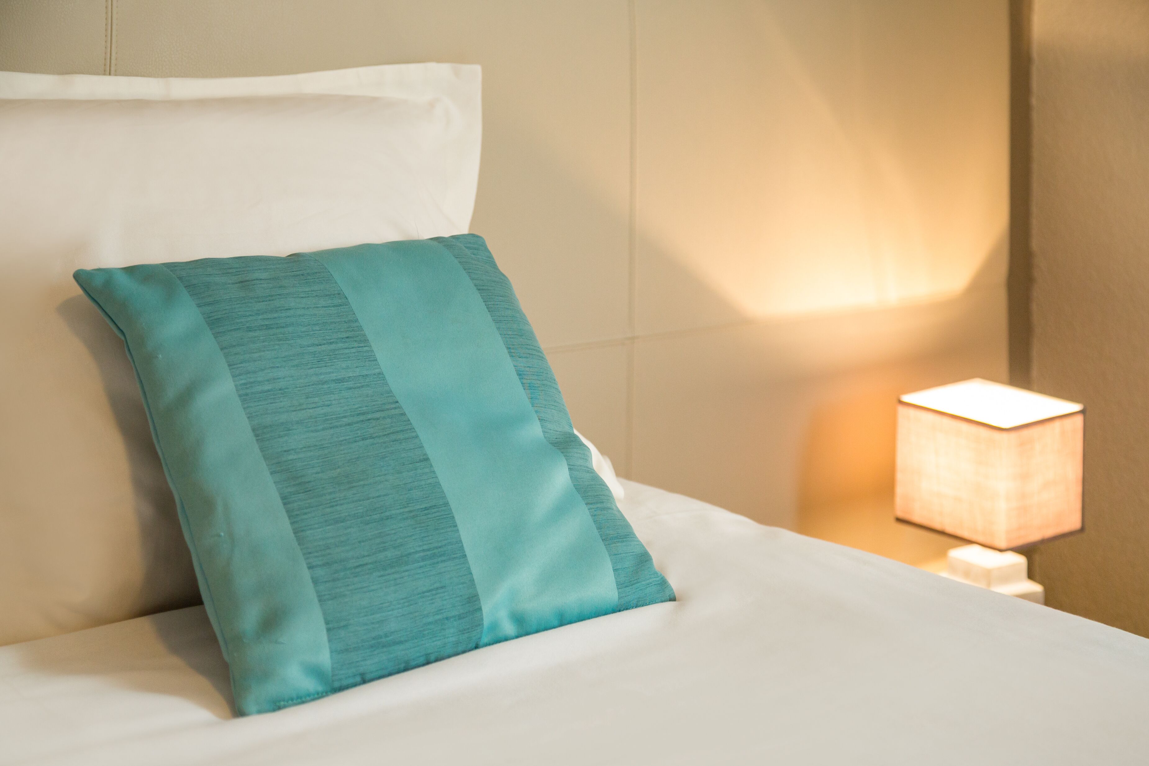 A neatly made bed with a teal pillow and a white pillow, a bedside lamp, and a wall with a textured finish.