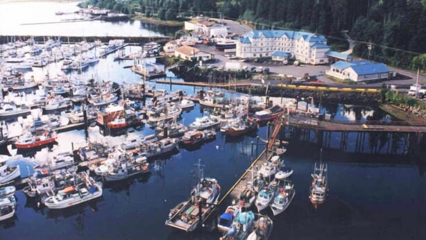 A marina with numerous boats docked, a large building with a blue roof, and a forested area in the background. - Quarterdeck Inn Port Hardy (Port Hardy)