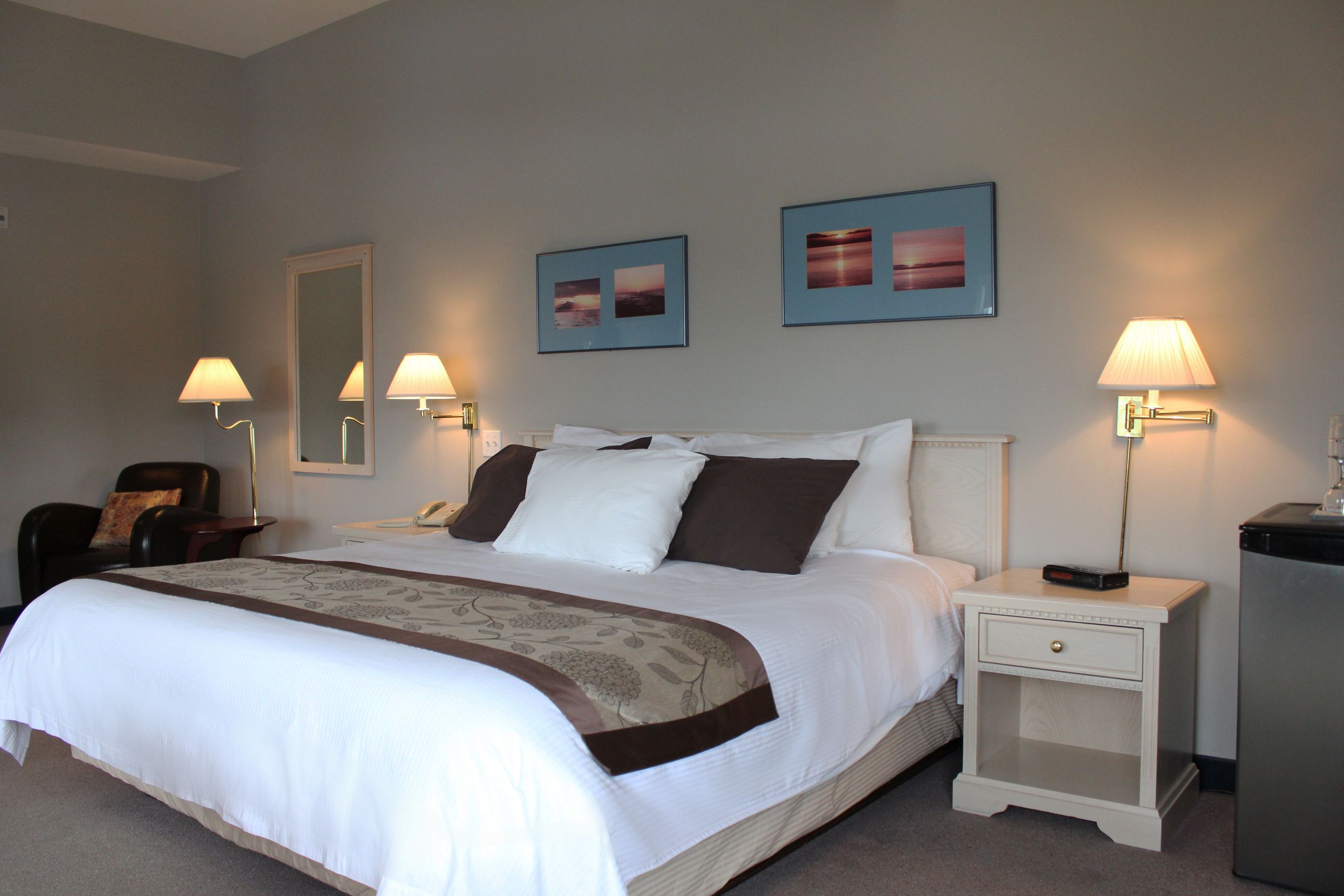 A neatly made bed with a white and brown bedspread, two bedside lamps, a nightstand with a drawer, and two framed pictures on the wall.
