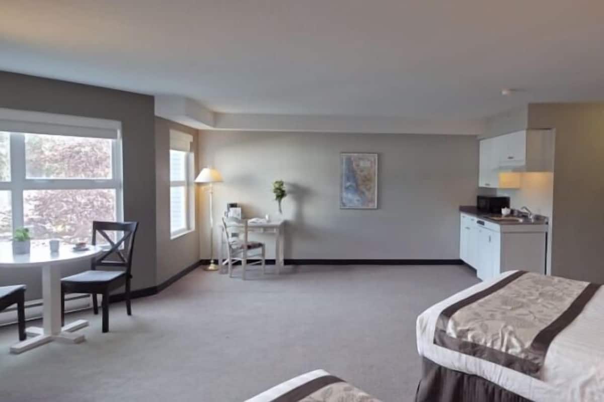 Quarto de hotel com cama, escrivaninha, cadeira, mesa pequena, janela, pintura e cozinha compacta.