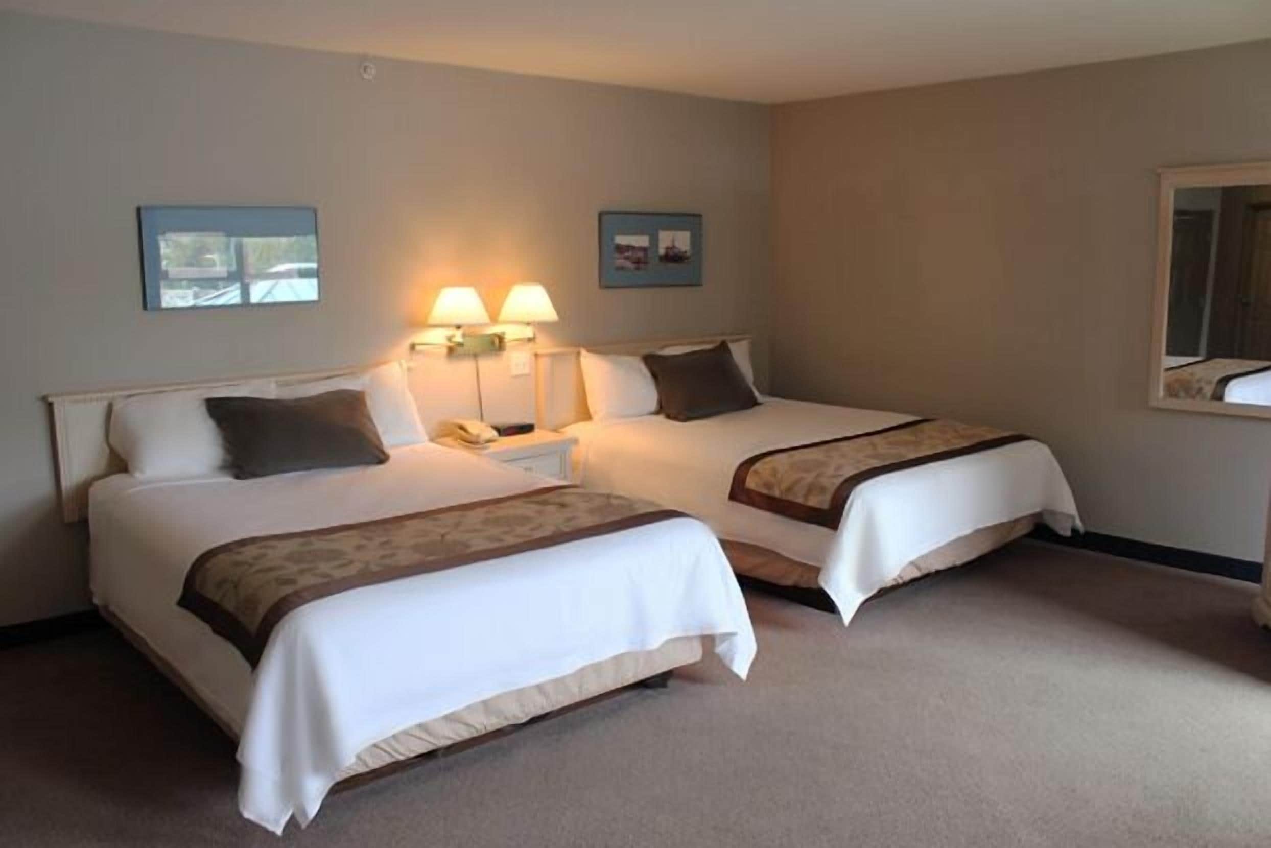 Une chambre d’hôtel avec deux lits, chacun agrémenté de draps blancs et d’un plaid marron. Deux lampes murales sont fixées de chaque côté des lits, et deux tableaux encadrés sont accrochés au-dessus.