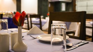 Une table à manger dressée avec un verre transparent, un vase blanc contenant une fleur rouge, des salières et des poivrières, et une assiette blanche.