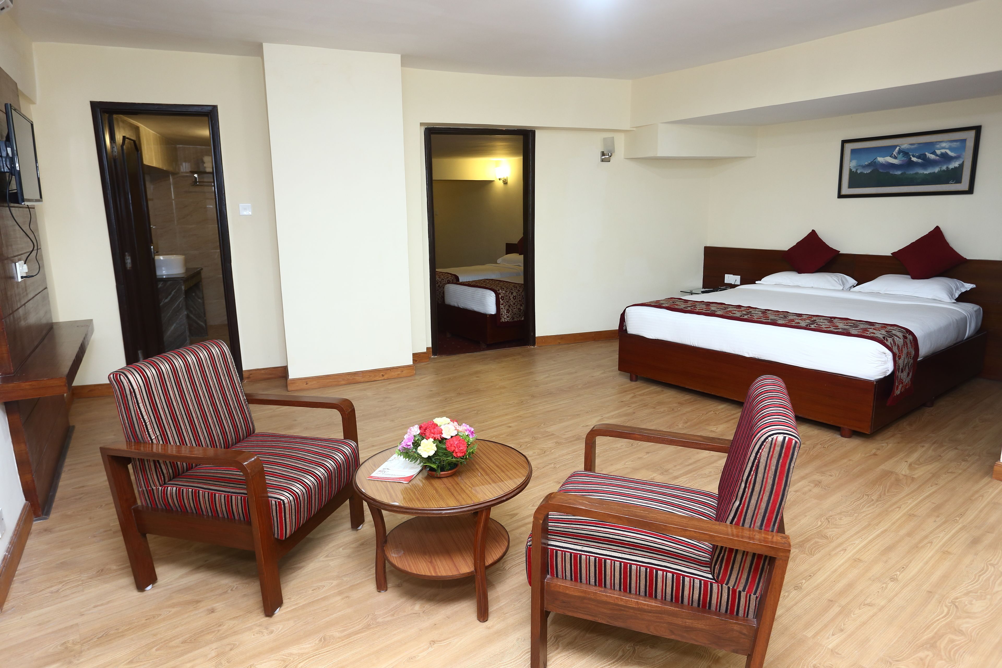 A hotel room with a large bed, two striped armchairs, a small wooden table with a vase of flowers, and a painting on the wall.