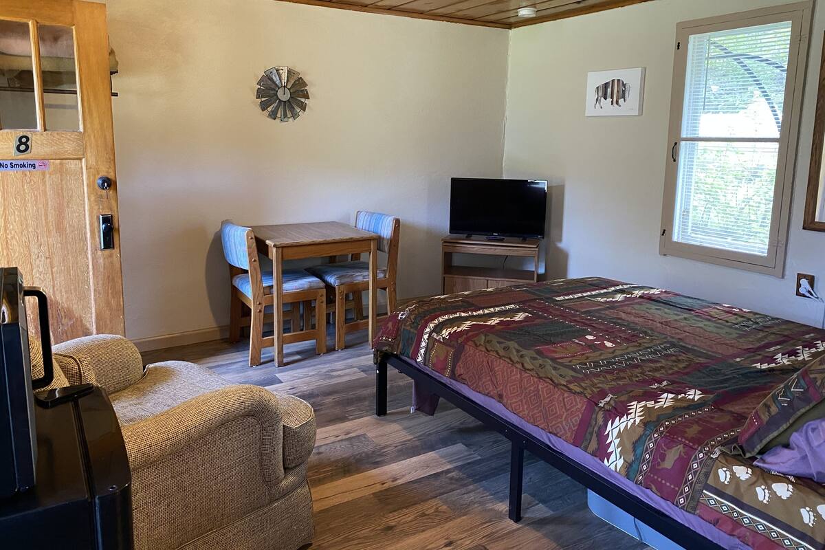 Una habitación con una cama, televisión, escritorio con dos sillas, un reloj en la pared y una ventana con persianas.