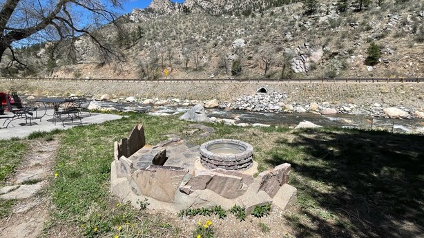 A riverside picnic area with a stone fire pit, a table and chairs, and a view of a mountain range. - Stoney River Lodge (Loveland)