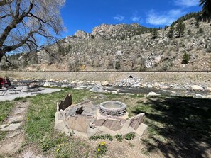 A riverside picnic area with a stone fire pit, a table and chairs, and a view of a mountain range. - Stoney River Lodge (Loveland)