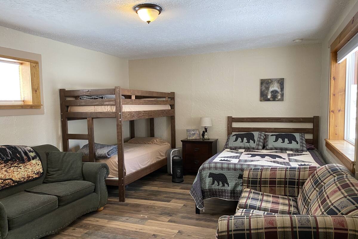 Una cama litera con cabecera de madera, un sofá verde, un sofá a cuadros, una mesita de noche con una lámpara y un cuadro colgado en la pared.