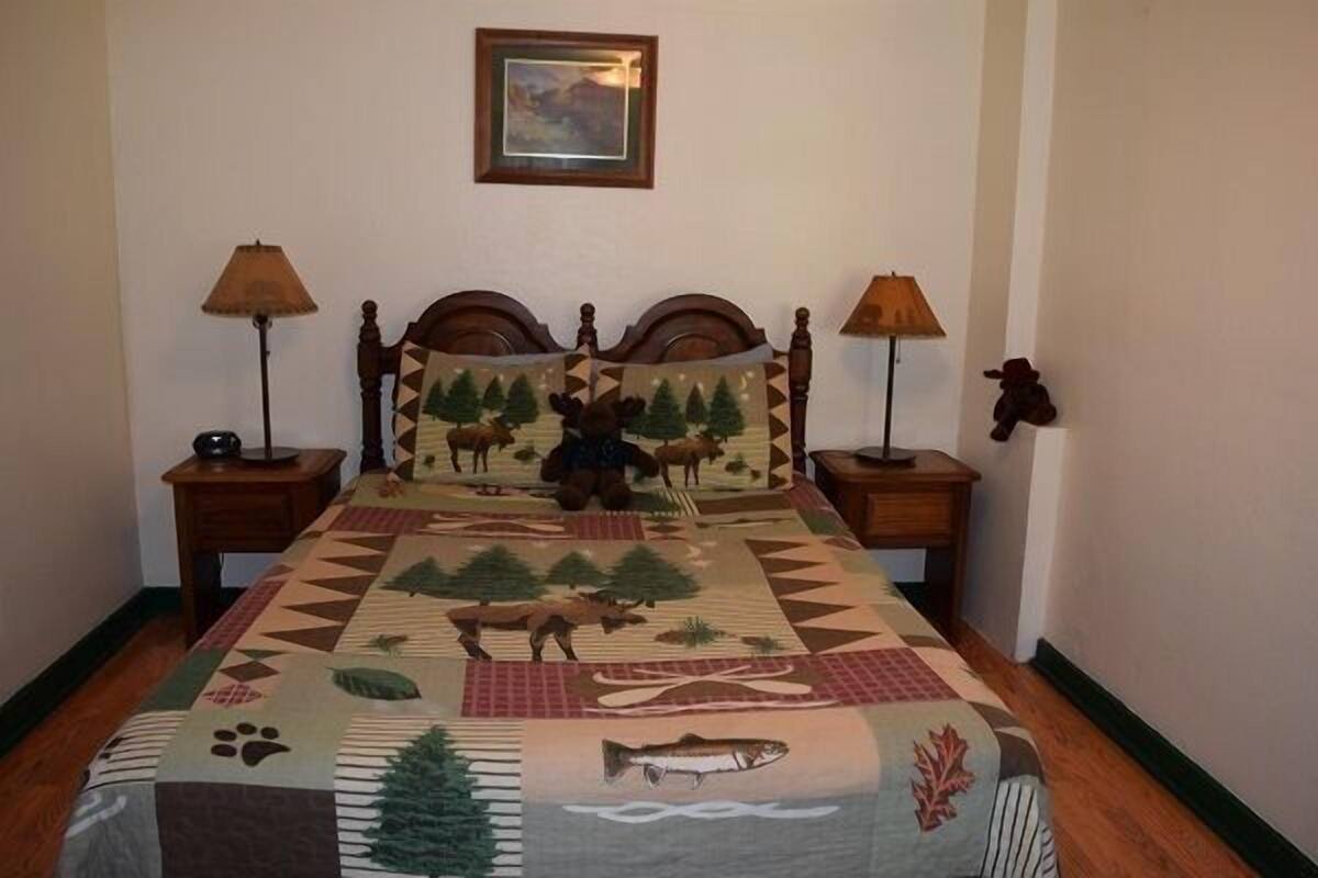 Una cama con colcha que muestra escenas de vida silvestre y paisajes naturales, flanqueada por dos lámparas de noche y una mesita con lámpara.