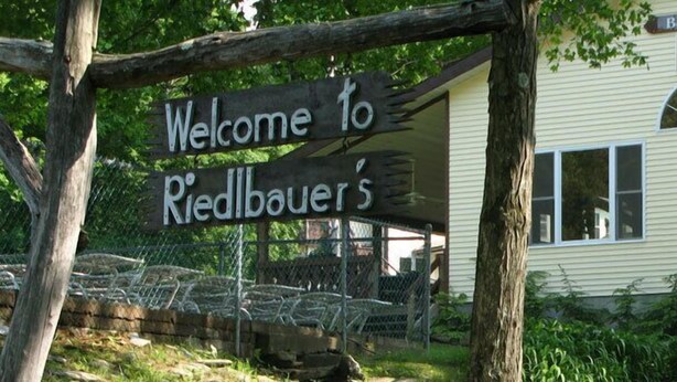 Un letrero de madera con la inscripción 'Welcome to Riedlbauer's', una casa con ventana y un árbol al fondo.