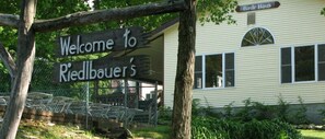 A wooden sign with "Welcome to Riedlbauer's" written on it, a house with a window, and a tree in the background.