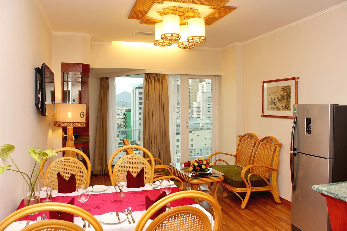 Een eetgedeelte met een tafel voor vier personen, omringd door rieten stoelen, een bijzettafeltje met een vaas, en uitzicht op de stad door grote ramen.