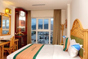 Habitación de hotel con una cama grande, un escritorio con silla, un espejo y vistas a la ciudad a través del ventanal.