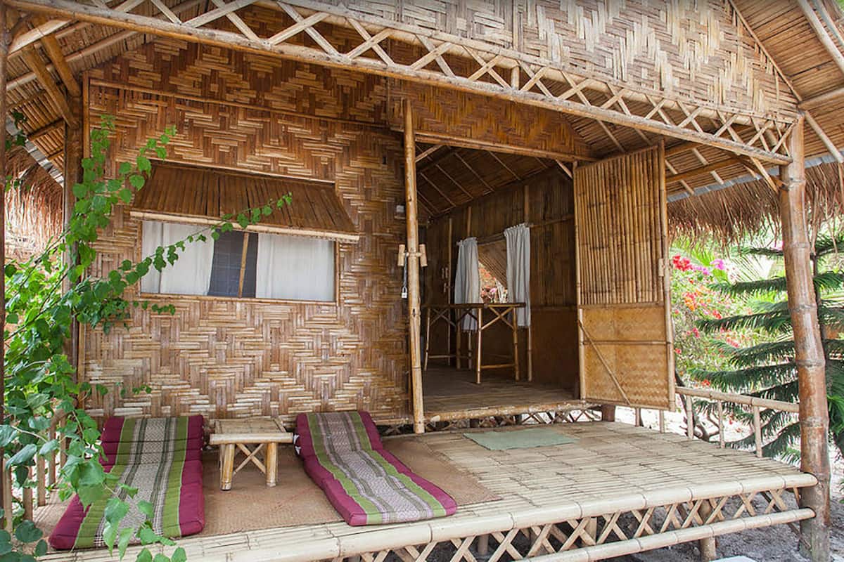 A bamboo structure with a thatched roof, a small enclosed area with a window, and a bench with cushions.