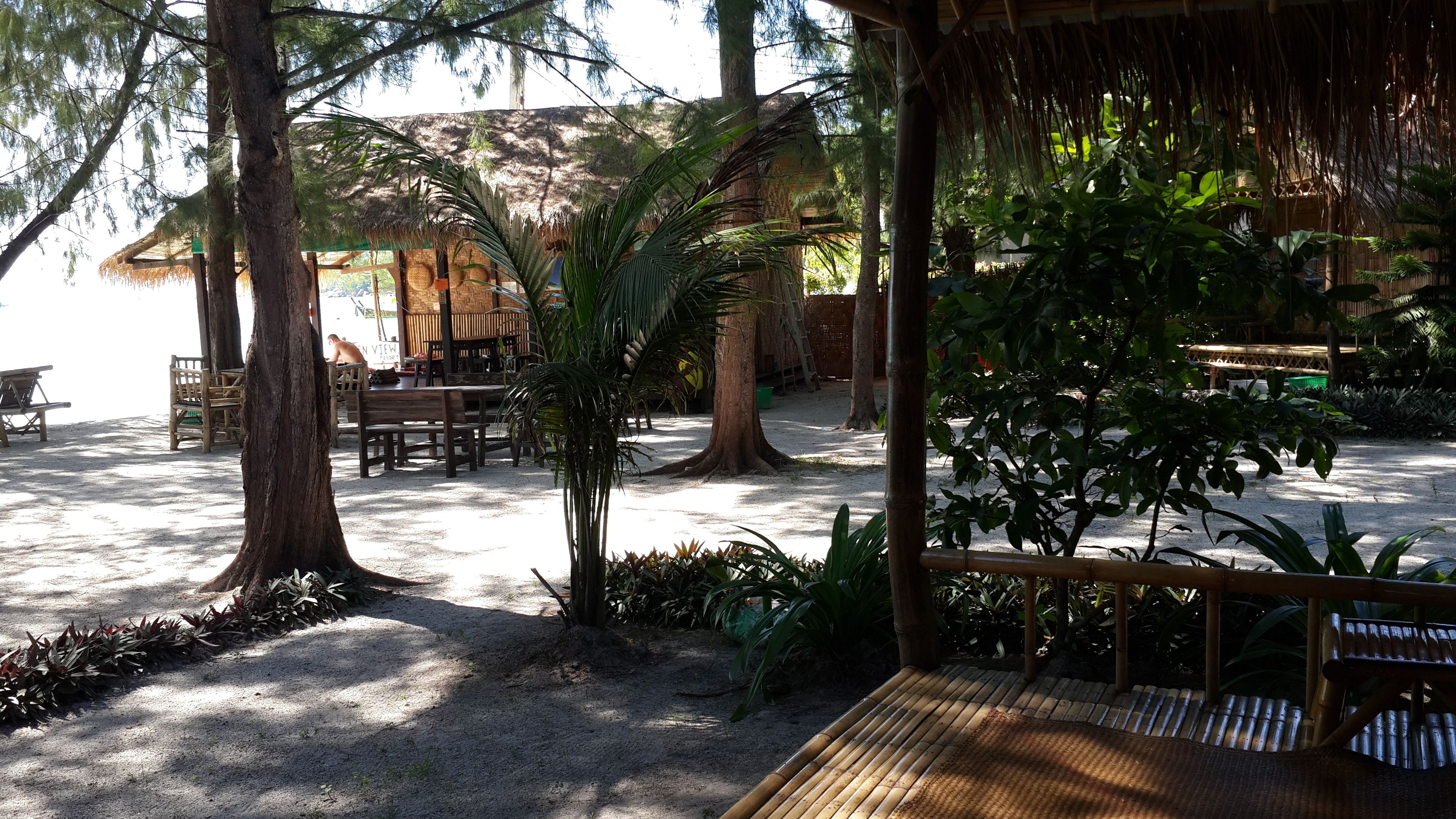 An outdoor seating area with wooden tables and chairs, surrounded by trees and a thatched-roof structure.