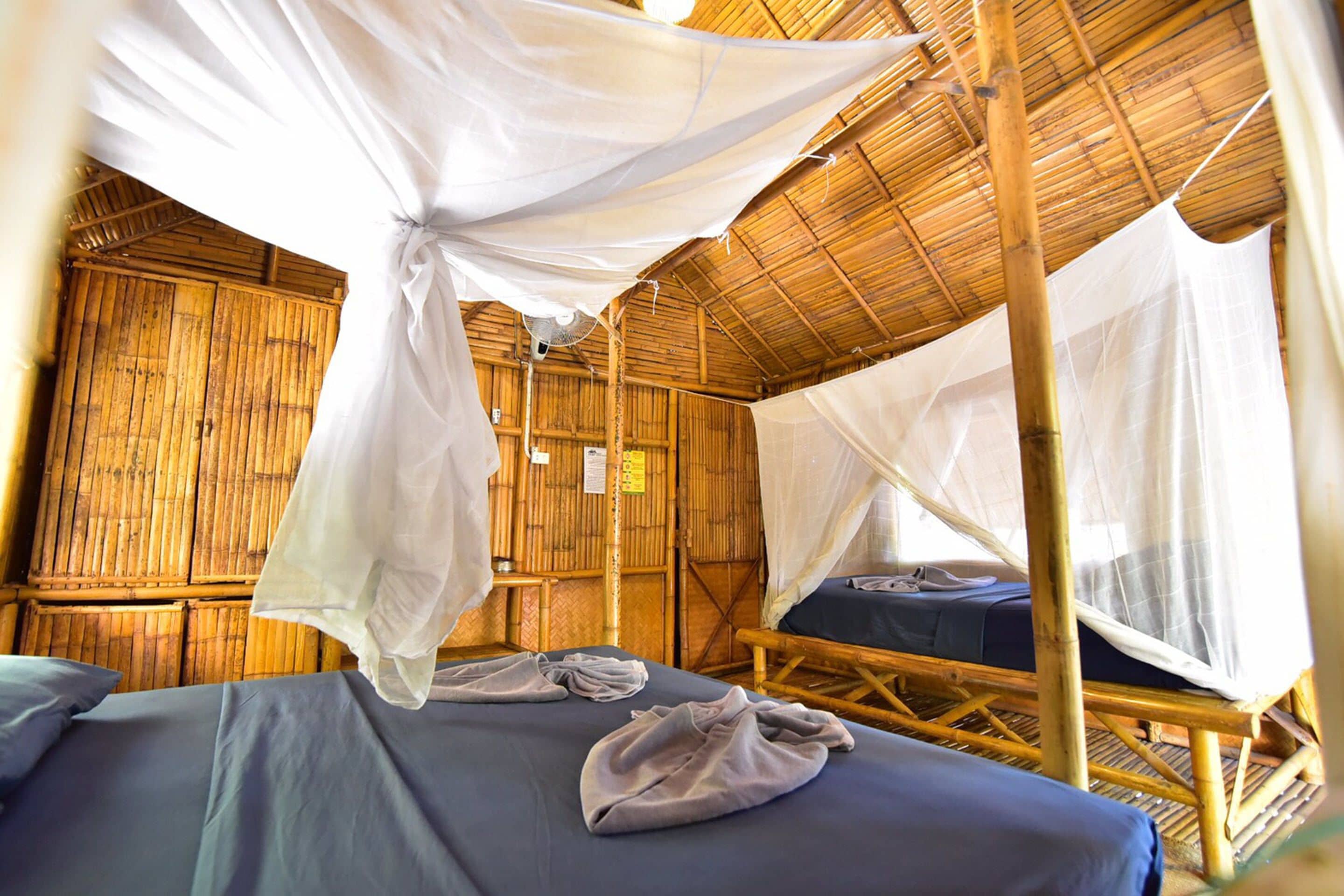 Una habitación con dos camas, cada una cubierta con mosquiteros y ropa de cama azul. La habitación tiene techo y paredes de bambú.