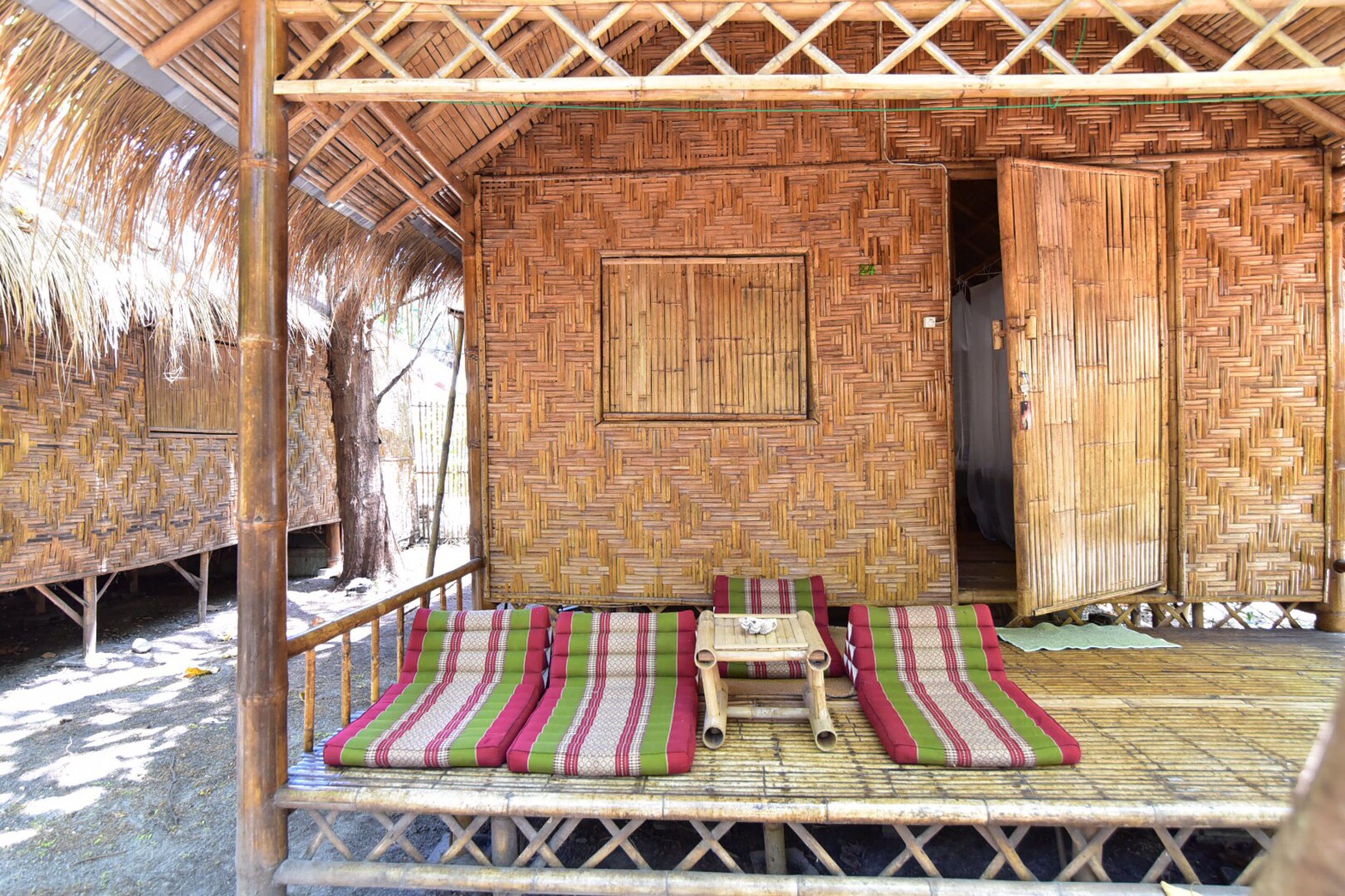 A bamboo hut with a thatched roof, a central bed with striped bedding, and a small table with a cup.