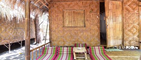 A bamboo hut with a thatched roof, a central bed with striped bedding, and a small table with a cup.
