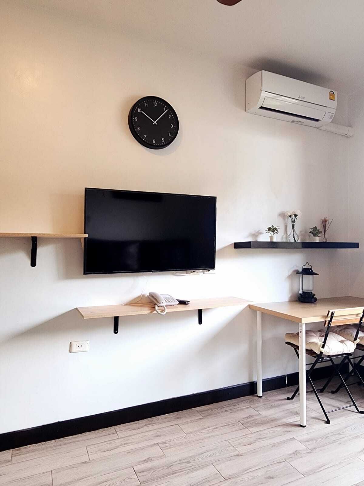 A modern living room with a mounted TV, a clock, an air conditioner, a small table with a lamp, and a chair.
