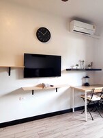 A modern living room with a mounted TV, a clock, an air conditioner, a small table with a lamp, and a chair.