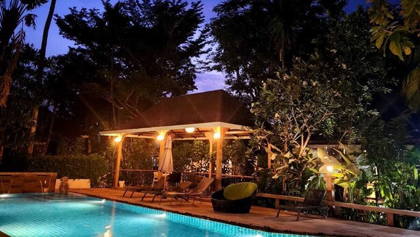A swimming pool with lounge chairs and a covered seating area, surrounded by trees and a clear sky. - Cocoville Phuket Resort (Chalong)