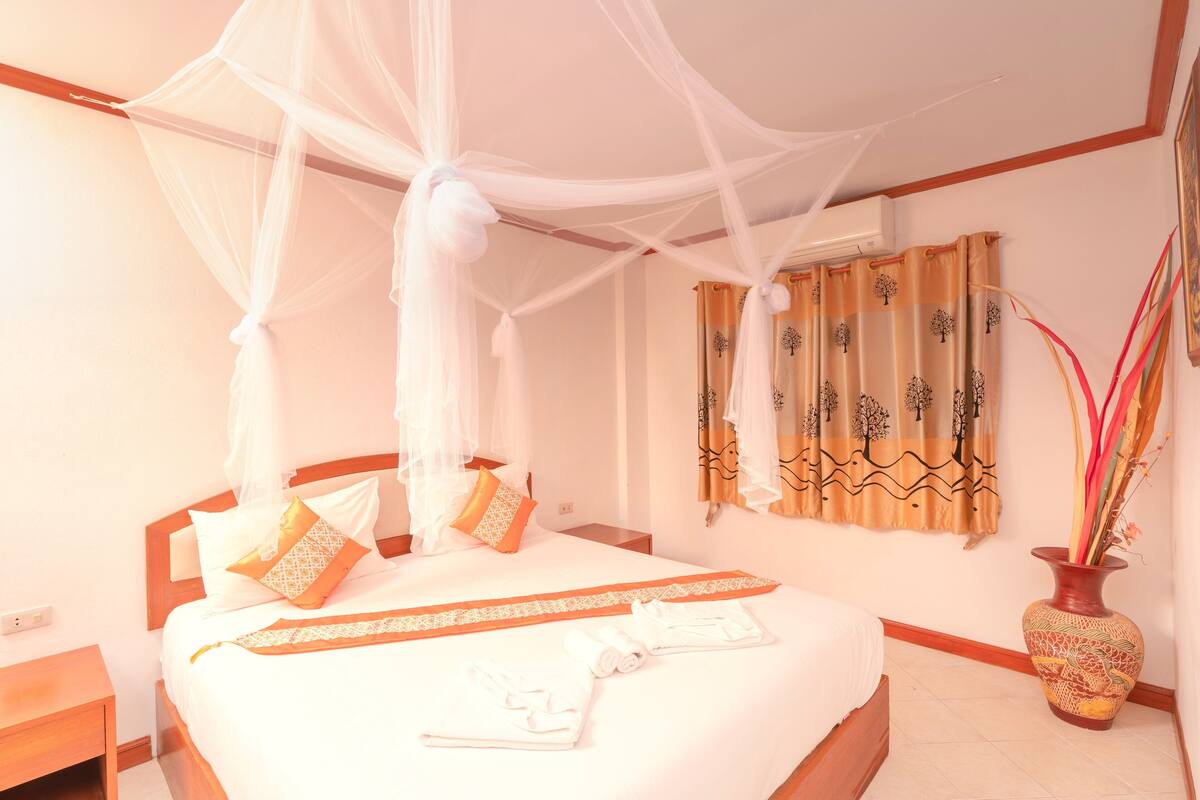A neatly made bed with a canopy, a vase with plants, and a wall with patterned curtains.
